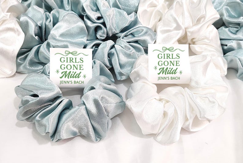 May include: Satin scrunchies in pale blue and white. Some scrunchies have a small white card with the text "GIRLS GONE Mild" and "Jenn's Bach" in green. The scrunchies are arranged on a white surface, ideal for a hen party.