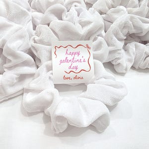 May include: A collection of white velvet scrunchies, with a small card that reads "happy galentine's day" in pink script, and "love, olivia" below. The scrunchies are gathered together, creating a soft, textured appearance.