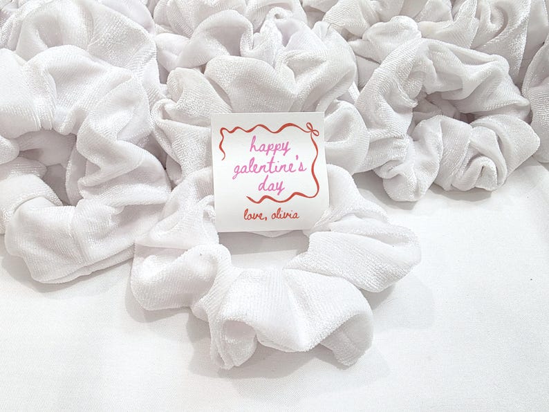 May include: A collection of white velvet scrunchies, with a small card that reads "happy galentine's day" in pink script, and "love, olivia" below. The scrunchies are gathered together, creating a soft, textured appearance.