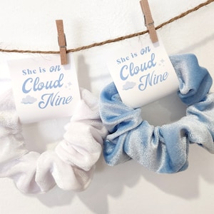 May include: Two velvet scrunchies, one white and one light blue, with a tag attached to each that reads "She is on Cloud Nine".