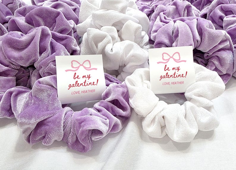 May include: A collection of soft scrunchies in shades of lavender and white. Each scrunchie is made of a plush material. Some scrunchies have a small card that reads "be my galentine!" in red script, with a pink bow graphic.