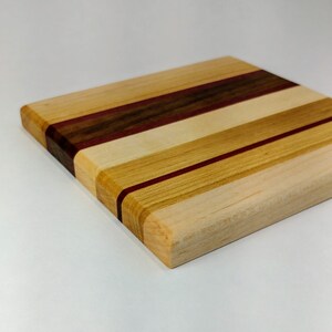May include: A square wooden cutting board with a multi-colored striped design. The board is made of maple, walnut, and purpleheart wood.