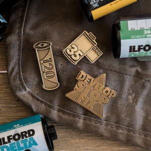 May include: Four gold-coloured metal pins with a film photography theme. The pins feature the text "DEVELOP STOP FIX", "120", "35", and a film canister.
