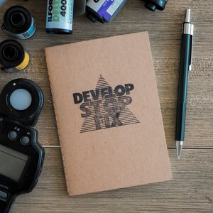 May include: A brown notebook with the words "DEVELOP STOP FIX" printed in black on the front. The notebook is sitting on a wooden surface with a black pen and a roll of film.