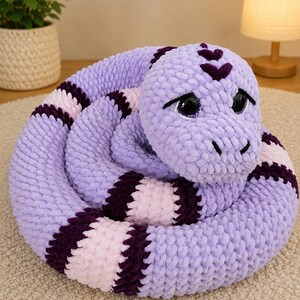 May include: A coiled plush snake toy crafted from chunky lavender, light pink, and dark purple yarn. The snake features large, glossy black eyes with purple glitter accents, a rounded head, and a textured, soft-touch knit surface throughout its body.