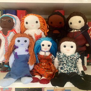 May include: A collection of eight handmade dolls, each with distinctive features and attire. The dolls are displayed on a white shelf with books behind them. The dolls are crafted from fabric and have a variety of hair and eye colours. Some of the dolls are wearing dresses, while others are wearing trousers and shirts.