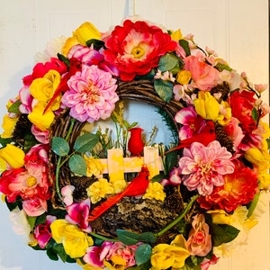 May include: A spring-themed wreath with a variety of yellow, pink, and red flowers. Two red cardinals are perched on a wooden fence with a yellow and white picket design. The wreath is made of woven twigs and has a rustic look.