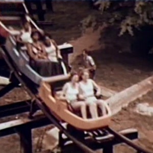 May include: A wooden roller coaster car with four people riding. The car is brown and yellow with a blue stripe. The people are wearing casual clothing and are smiling.
