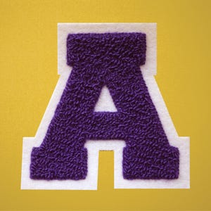 May include: A purple chenille letter "A" patch with a white felt border, set against a yellow background. The letter has a textured, raised appearance, commonly used for varsity jackets or personalized apparel.
