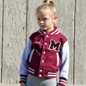 May include: A maroon and gray letterman jacket with the letter "M" on the front. The jacket has white stripes on the collar, cuffs, and hem. The name "Leah" is embroidered on the chest. The jacket is buttoned up.