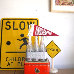 Könnte beinhalten: Ein rot-weißes Wimpel mit dem Wort "KINDERGARTEN" darauf hängt über einem Sechserpack Glasflaschen in einem Metallträger. Die Flaschen sind mit "Pepsi Cola" beschriftet. Im Hintergrund ist ein gelbes Schild mit den Worten "SLOW CHILDREN AT PLAY" zu sehen. Ein weiteres gelbes Schild mit dem Wort "SCHOOL" ist ebenfalls sichtbar.