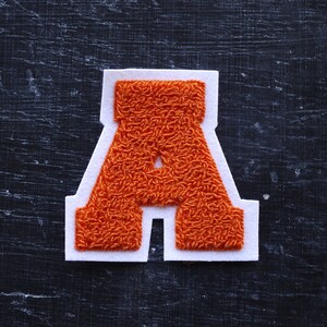 May include: Orange felt letter A with white outline. The letter is stitched with a fuzzy texture.