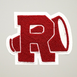 Puede incluir: Un parche bordado rojo con la forma de la letra "R" con un diseño de megáfono. La letra y el megáfono son rojos con un borde blanco. El parche tiene un aspecto texturizado.