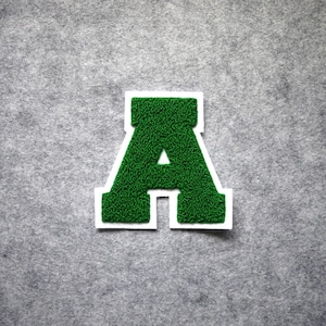 May include: A green letter "A" patch with a white border, set against a gray background. The letter has a textured, fuzzy appearance. This patch is suitable for clothing or accessories.