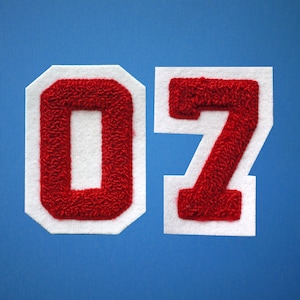 Op de afbeelding: Twee witte viltpleisters met rode viltcijfers 0 en 7. De cijfers zijn in een klassiek lettertype in de stijl van een sporttrui.