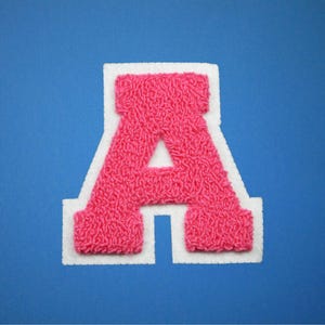 May include: A pink chenille letter "A" patch with a white border, set against a blue background. The letter has a textured, fuzzy appearance, and the white border provides a clean contrast. This patch is suitable for various craft projects.