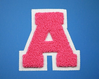 May include: A pink chenille letter "A" patch with a white border, set against a blue background. The letter has a textured, fuzzy appearance, and the white border provides a clean contrast. This patch is suitable for various craft projects.