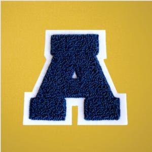 May include: A navy blue chenille letter "A" patch with a white border, set against a yellow background. The letter has a textured, raised appearance, typical of varsity-style patches. Ideal for jackets, bags, or other fabric items.