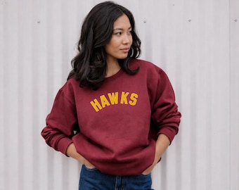 May include: A maroon sweatshirt with the word "HAWKS" in yellow block letters. The sweatshirt is worn by a person with dark hair and blue jeans. The background is a white corrugated metal wall.