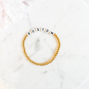 May include: Gold beaded bracelet with white letter beads spelling out the word "BOSTON".