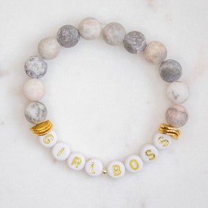 May include: A gray and white beaded bracelet with gold accents and white letter beads spelling "GIRL BOSS".