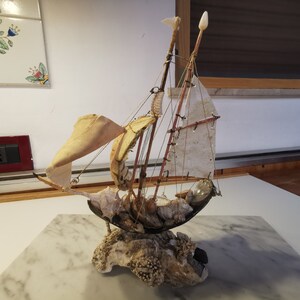 Op de afbeelding: Een klein, handgemaakt model van een zeilboot gemaakt van schelpen en andere natuurlijke materialen. De zeilen zijn van witte stof en de romp is gemaakt van een grote schelp. De boot staat op een basis van koraal en andere schelpen.