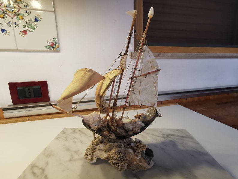 Op de afbeelding: Een klein, handgemaakt model van een zeilboot gemaakt van schelpen en andere natuurlijke materialen. De zeilen zijn van witte stof en de romp is gemaakt van een grote schelp. De boot staat op een basis van koraal en andere schelpen.