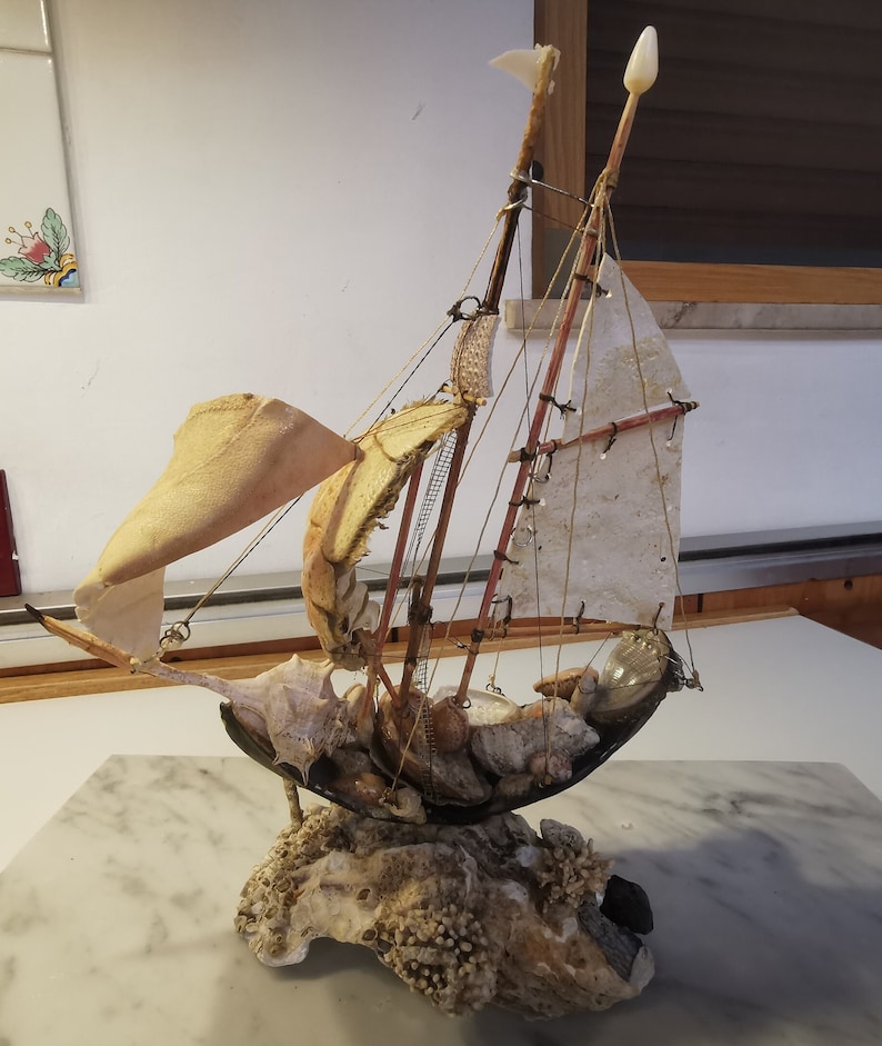 Op de afbeelding: Een miniatuur zeilboot gemaakt van schelpen en andere natuurlijke materialen. De boot heeft een wit zeil en een bruine houten mast. De boot staat op een basis van koraal en schelpen.