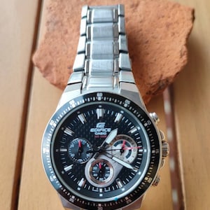 May include: A silver and black chronograph wristwatch with a black face and red accents. The watch has a silver metal band and the words "EDIFICE CASIO" on the face.