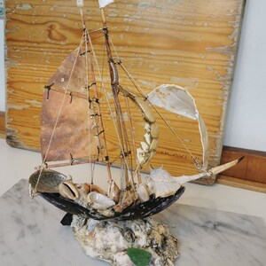 Op de afbeelding: Een miniatuur zeilboot gemaakt van schelpen en drijfhout. Het zeil is van bruine stof en de mast is van een dun stuk hout. De boot staat op een basis van schelpen en drijfhout.