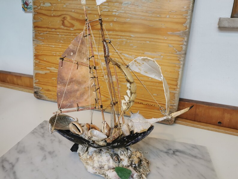 Op de afbeelding: Een miniatuur zeilboot gemaakt van schelpen en drijfhout. Het zeil is van bruine stof en de mast is van een dun stuk hout. De boot staat op een basis van schelpen en drijfhout.