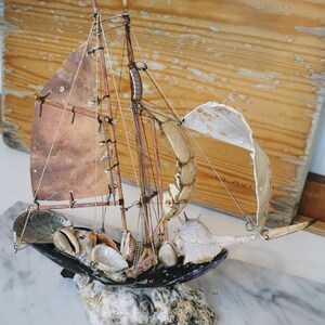 Op de afbeelding: Een miniatuur zeilboot gemaakt van schelpen en drijfhout. De boot heeft een wit zeil en een bruine romp. Het is versierd met verschillende schelpen, waaronder een grote schelp en een kleinere schelp. De boot staat op een bed van schelpen en drijfhout.