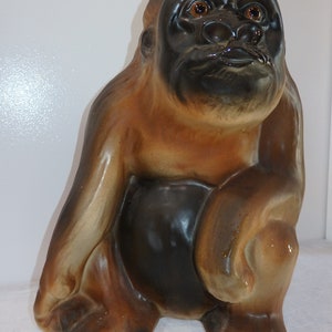 May include: A ceramic gorilla ornament in shades of brown and black. The gorilla is depicted in a seated pose, with one arm resting on its leg. The figurine has detailed facial features and a glossy finish.