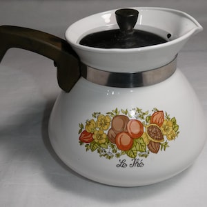 May include: White ceramic teapot with a black lid and a dark green handle. The teapot has a silver band around the middle and a floral design with the text "Le Thé".