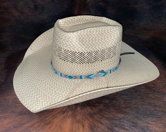 May include: A light beige straw cowboy hat featuring a woven chevron pattern and a ventilated band around the crown. The hat is accented with a thin, decorative beaded hatband in shades of blue, grey, and white, featuring a geometric diamond motif.