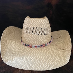 May include: A straw cowboy hat with a beaded band featuring the American flag colors and the word "USA" repeated. The hat is beige and has a woven pattern.