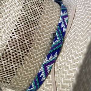 May include: A light tan woven straw hat featuring a decorative band made of small beads in a geometric pattern. The beadwork displays a repeating design in shades of royal blue, purple, white, and turquoise, secured with a thin black cord.