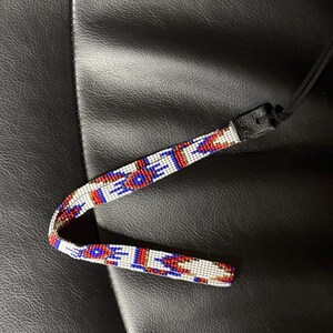 May include: A beaded strap with a geometric pattern in red, white, and blue. The strap has a black leather end and a black cord. The beaded section is approximately 0.5 inches wide.