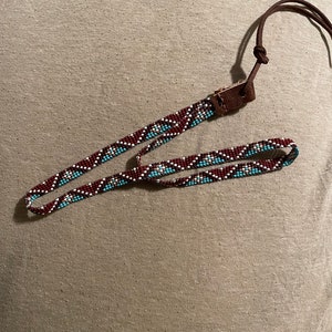 May include: A long, thin beaded strap featuring a geometric pattern in dark red, light blue, and white glass beads. The strap is finished with a brown leather connector and a thin brown leather cord tied in a knot at the end of the assembly.