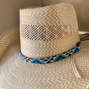 May include: A light beige woven straw hat featuring a wide brim and a tall crown with a ventilated section. A decorative band of small glass beads in geometric patterns of black, white, and shades of blue encircles the base of the crown.