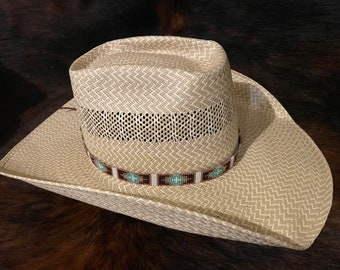 May include: A light beige straw cowboy hat featuring a woven chevron pattern, a ventilated mid-section, and a decorative hat band adorned with a geometric beadwork design in shades of brown, white, and turquoise blue around the base.