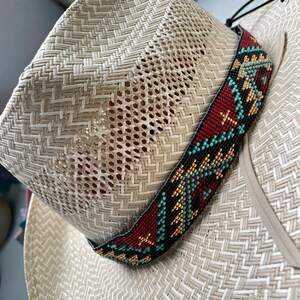 May include: A light beige woven straw hat featuring a wide band decorated with intricate beadwork. The beadwork displays a geometric pattern in shades of deep red, turquoise, and metallic gold, encircling the base of the hat crown.