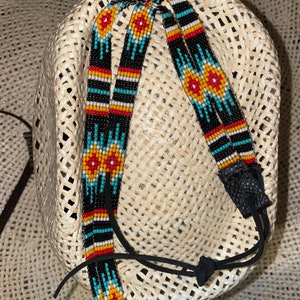 May include: A hat band featuring intricate beadwork in black, white, red, yellow, and turquoise. The design includes geometric patterns and floral-like motifs. The band is finished with black leather tabs and a black cord with a knot for adjustment.