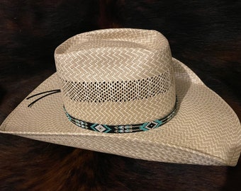 May include: A light beige straw cowboy hat featuring a woven texture, a ventilated crown section, and a decorative hat band adorned with a geometric pattern in black, white, and turquoise seed beads, finished with two thin black leather cords.