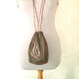 Peut inclure: Sac en cuir vert olive avec cordon de serrage et motif en forme de flamme en cuir couleur cuivre. Le sac est suspendu à une longue et fine lanière en cuir marron. Le sac est présenté sur un mannequin.