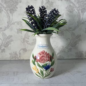 May include: A small white ceramic vase with a floral design. The vase is hand-painted with pink, yellow, blue, and green flowers. The vase has a label that says "Handpainted" and "American Creek Pottery".