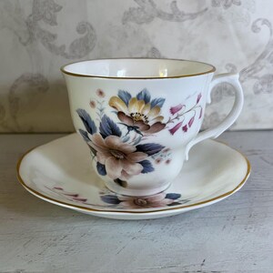 May include: A white teacup and saucer with a floral pattern in shades of pink, blue, and yellow. The cup has a gold rim and the saucer has a gold band around the edge.
