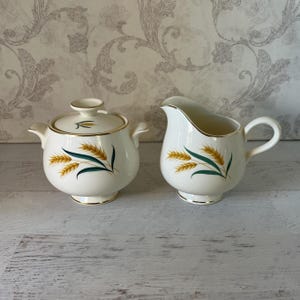 May include: A white sugar bowl and creamer set with a gold trim and a wheat design. The set is made of ceramic and has a vintage look.