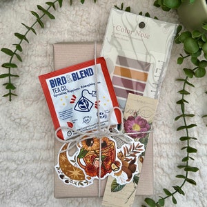 Puede incluir: Una caja de regalo llena de una bolsita de té, un marcapáginas y una pegatina. La bolsita de té es de Bird & Blend Tea Co. y dice "Spreading Happiness. Reimagining ca." El marcapáginas es de estilo vintage con un diseño floral. La pegatina es una ilustración colorida de una taza de té con girasoles y estrellas.