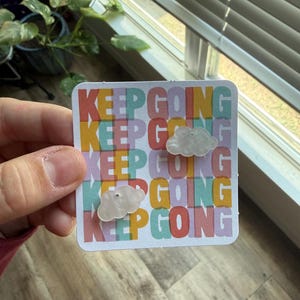 May include: A pair of white, cloud-shaped stud earrings attached to a square display card. The card features the repeating text KEEP GOING in a multi-coloured, layered font with shades of red, yellow, blue, and green against a white background.
